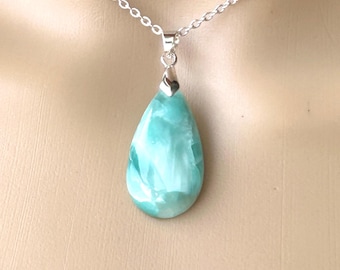 May include: A teardrop-shaped pendant necklace featuring a large, light blue gemstone with white marbling. The pendant hangs from a delicate silver chain. The gemstone has a polished, smooth surface.