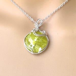 May include: A round, lime green, mottled gemstone pendant encased in a silver-colored wire-wrapped setting. The pendant hangs from a delicate silver-colored metal chain. The stone features a smooth, polished surface with visible internal texture.