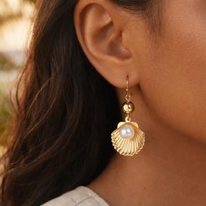 Peut inclure: Une boucle d'oreille pendante dorée composée d'une breloque en forme de coquillage en métal poli suspendue à une petite sphère dorée. Une perle blanche est fixée au centre du coquillage, qui est accroché à un crochet d'oreille simple.