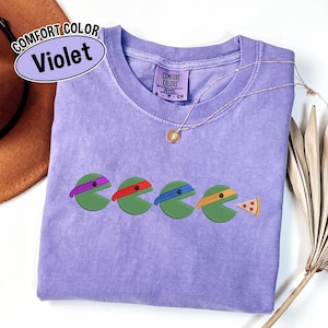 May include: A violet Comfort Colors t-shirt featuring an embroidered graphic of four green circular characters with purple, red, blue, and orange eye masks, with the last character eating a slice of pizza. The shirt has a crew neck and a size S label.