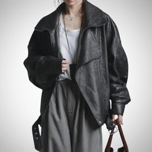 May include: A black leather jacket with a wide collar and a belted waist. The jacket has a slightly oversized fit and a textured appearance. The model is wearing a white shirt and gray pants.