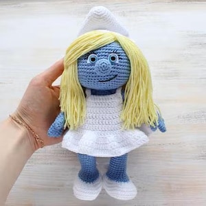 Könnte beinhalten: Handgefertigte Häkelpuppe der Figur Schlumpfine. Die Puppe hat blaue Haut, gelbes Haar und trägt ein weißes Kleid und einen Hut. Die Puppe ist etwa 20 cm groß.