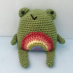 May include: A green crochet frog toy with a rainbow belly. The frog has small black eyes and a stitched mouth. A handmade soft toy, perfect for children or as a decorative item.