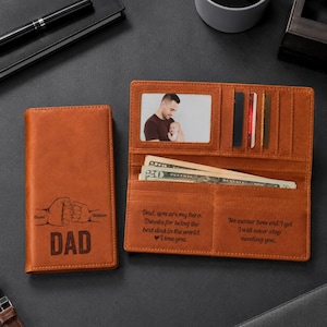 May include: A brown leather wallet, closed and open, with a photo and cash visible. The closed wallet has the word "DAD" and two fists engraved. The open wallet has text that reads "Dad, you are my hero..." and "No matter how old I get..."
