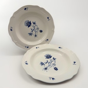May include: Two off-white ceramic plates with scalloped edges, each featuring a central dark blue floral pattern and small blue flower motifs scattered around the rim. The plates have a smooth, glossy finish and a rounded, slightly raised border.