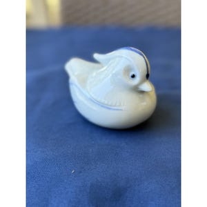 May include: A small white ceramic figurine of a duck with a smooth, glossy finish. The duck features delicate blue painted accents along its head, back, and wings, with a single dark blue dot marking the eye and a small crest on its head.