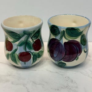 May include: Two ceramic mugs with a cream-colored base and a light blue vertical stripe pattern. The left mug features hand-painted red cherries with green leaves, while the right mug displays large, dark purple plums with green foliage.