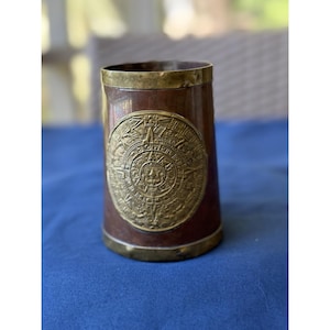 May include: A copper-colored metal cup with a brass rim and a large, detailed brass Aztec calendar design. The cup has a cylindrical shape and a slightly flared base, showcasing a blend of textures and colors.