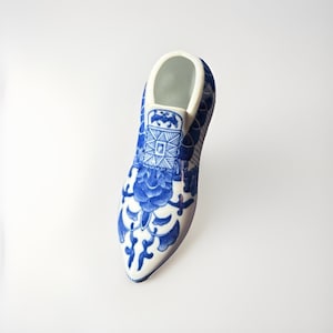 May include: A porcelain shoe figurine in white and blue. The shoe features a pointed toe and a high heel. The blue floral pattern covers the shoe's surface, with a white interior and trim. The shoe is a decorative item.