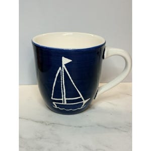 May include: A dark blue ceramic mug featuring a white hand-painted sailboat illustration on the front. The mug has a smooth, glossy finish, a white interior, and a white handle. The sailboat design shows a mast, sails, and a hull.