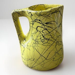 May include: A yellow ceramic pitcher with a black scribble design. The pitcher has a unique handle and a square-shaped opening. The design is a modern, abstract style, suitable for serving beverages or as a decorative piece.