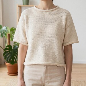 May include: A short-sleeved, cream-colored knit sweater featuring a round neckline with a rolled edge, drop shoulders, and a straight hemline. The garment is constructed with a visible stockinette stitch pattern and has a relaxed, boxy fit.