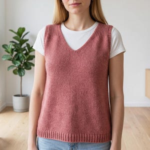 Könnte beinhalten: Eine altrosa Strickweste mit V-Ausschnitt und gerippten Abschlüssen an den Armausschnitten, am Ausschnitt und am Saum, getragen über einem schlichten weißen kurzärmeligen T-Shirt und einer hellblauen Jeans, die einen einfachen, klaren Lagenlook zeigt.