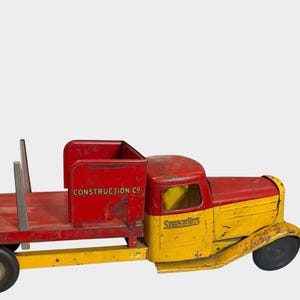 May include: A vintage metal toy truck with a red flatbed and a yellow cab. The truck features a red side panel with the text CONSTRUCTION CO and a yellow door with the text STRUCTO TOYS. It has six vertical metal stakes on the flatbed and black wheels.