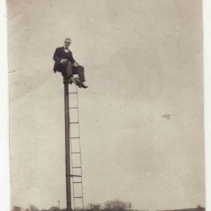 May include: A black and white photo of a man sitting on a ladder attached to a tall pole. The pole is near a set of railroad tracks.