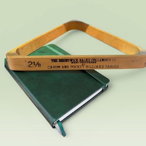 May include: A triangular wooden billiard ball rack with the text 'THE BRUNSWICK BALKE COLLENDER CO MANUFACTURERS OF CAROM AND POCKET BILLIARD TABLES' and '2 1/8' printed on the side, resting on a dark green rectangular notebook with an elastic band.