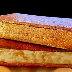 May include: Two antique books with worn, textured leather covers. The top book is a light brown color, and the bottom book is a darker reddish-brown. The pages are aged, with a yellowed appearance. The books are stacked on top of each other.