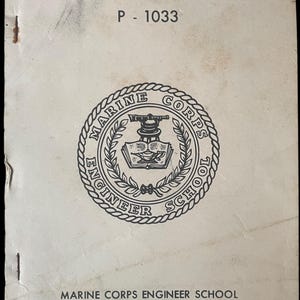 Puede incluir: Un "Manual del Electricista" vintage de la Escuela de Ingenieros del Cuerpo de Marines, P-1033. La portada presenta un emblema circular con texto y una ilustración detallada. El manual es de Camp Lejeune, Carolina del Norte.