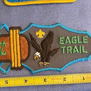 May include: A brown and gray embroidered patch with a blue border. It features a stylized eagle with a yellow scout symbol and the text "Eagle Trail".