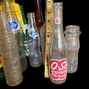 May include: A collection of vintage soda bottles, including a clear bottle with a red and white "O-So Beverages" label. The bottles vary in shape, size, and color, with a ruler showing the height of one bottle to be approximately 8 inches.