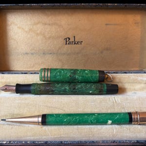 May include: An open vintage pen case with a green and black marbled fountain pen, a pen cap, and a matching pencil. The case is lined with beige velvet and has the "Parker" logo on the inside of the lid.