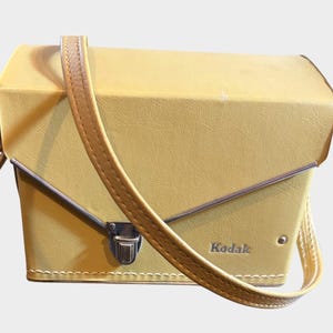 May include: A pale yellow rectangular camera case with a textured finish and a matching shoulder strap. The case features a silver-toned metal latch closure, a silver trim along the front flap, and the word Kodak embossed in the bottom right corner.