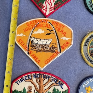 Puede incluir: Una colección de parches vintage de Boy Scouts que presentan diferentes nombres de senderos y ubicaciones. Los parches están bordados con diseños y texto coloridos, que incluyen "Tomahawk Trail", "Gateway West Historical Trail", "Three Notch Trail" y "Old Dominion Council BSA".