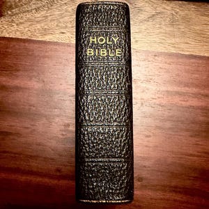 May include: A black, textured leather-bound book with the words HOLY BIBLE embossed in gold lettering on the spine. The spine features five horizontal decorative ridges, providing a classic look to this religious text, which is presented vertically.