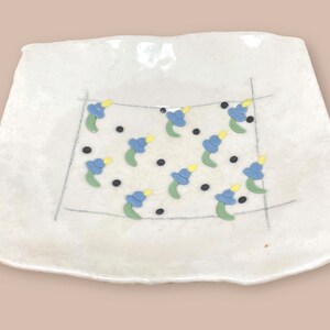 May include: A white ceramic square plate with a hand-painted design of blue, yellow, and green flowers. The plate has a slightly irregular edge.