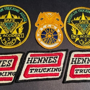 Könnte beinhalten: Fünf gestickte Aufnäher, drei sind rund und zwei rechteckig. Die runden Aufnäher sind goldfarben mit schwarzen Mittelpunkten und grünem Text. Der Text lautet "American Trucking Assns. Safe Driving Award No Accident 3 Years" und "American Trucking Assns. Safe Driving Award No Accident 2 Years". Die rechteckigen Aufnäher sind weiß mit rotem Text, der "Hennes Trucking" lautet.