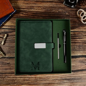 Puede incluir: Un juego de regalo verde oscuro que incluye un diario con cierre plateado, un bolígrafo y una recarga de bolígrafo. El diario está grabado con las iniciales "M" e "IVI" y el nombre "Michael".