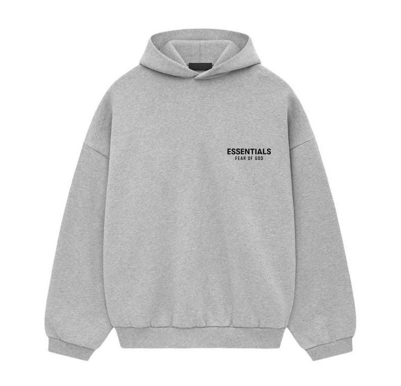 K&ouml;nnte beinhalten: Ein hellgrauer Kapuzenpullover mit Kapuze und langen &Auml;rmeln. Der Text "ESSENTIALS FEAR OF GOD" ist in Schwarz auf der oberen linken Brustseite aufgedruckt. Der Kapuzenpullover ist aus einem weichen, bequemen Material gefertigt.