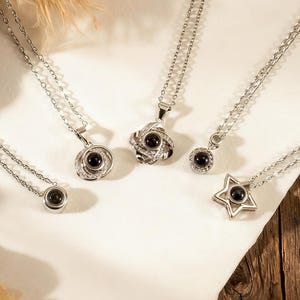 May include: Five silver necklaces with various pendant designs, including a circle, a flower, a star, and a round pendant. Each necklace has a small, black, circular stone. The necklaces are displayed on a white surface with a wooden background.