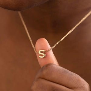 May include: A gold-colored necklace with a small, block letter "S" pendant. The pendant hangs from a delicate chain. The necklace is shown against a dark background.