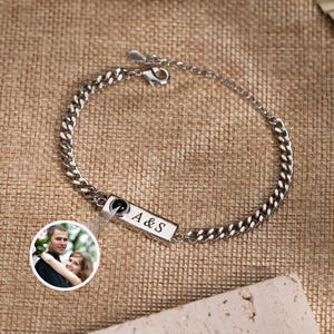 May include: Silver curb chain bracelet with a rectangular charm engraved with "A & S". The bracelet features a lobster clasp closure. A small, circular photo of a couple is attached to the bracelet.