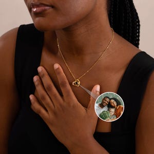 Peut inclure: Un collier pendentif en forme de c&oelig;ur dor&eacute; avec une petite pierre noire et des cristaux transparents. Le collier est port&eacute; autour du cou d'une personne. Une petite image circulaire d'une famille est dans le coin inf&eacute;rieur droit.