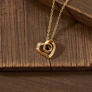 Peut inclure: Un collier pendentif en forme de c&oelig;ur dor&eacute; avec une cha&icirc;ne. Le pendentif comporte une pierre sombre au centre et est orn&eacute; de petites pierres claires. Le collier est pos&eacute; sur une surface en bois.