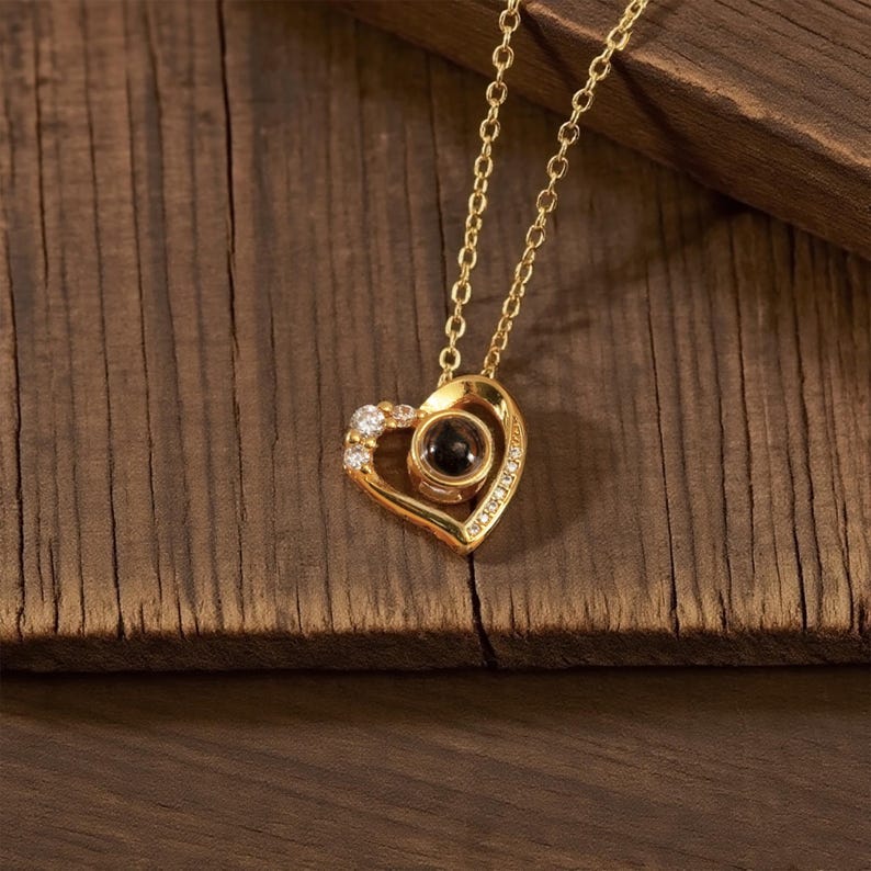 Peut inclure: Un collier pendentif en forme de c&oelig;ur dor&eacute; avec une cha&icirc;ne. Le pendentif comporte une pierre sombre au centre et est orn&eacute; de petites pierres claires. Le collier est pos&eacute; sur une surface en bois.