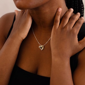 Peut inclure: Un collier dor&eacute; avec un pendentif en forme de c&oelig;ur. Le pendentif pr&eacute;sente une pierre noire au centre, entour&eacute;e de petites pierres transparentes. Le collier est port&eacute; autour du cou.