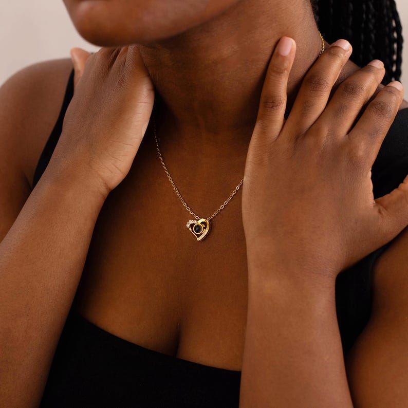 Peut inclure: Un collier dor&eacute; avec un pendentif en forme de c&oelig;ur. Le pendentif pr&eacute;sente une pierre noire au centre, entour&eacute;e de petites pierres transparentes. Le collier est port&eacute; autour du cou.