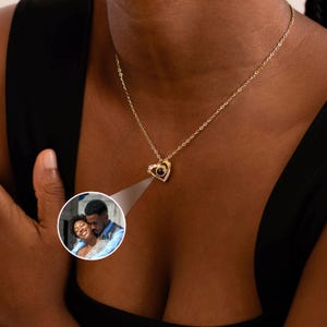 Peut inclure: Un collier pendentif en forme de c&oelig;ur dor&eacute; avec une petite pierre centrale ronde et sombre. Le pendentif est entour&eacute; de petites pierres claires. Une petite image ronde d'un couple est projet&eacute;e.