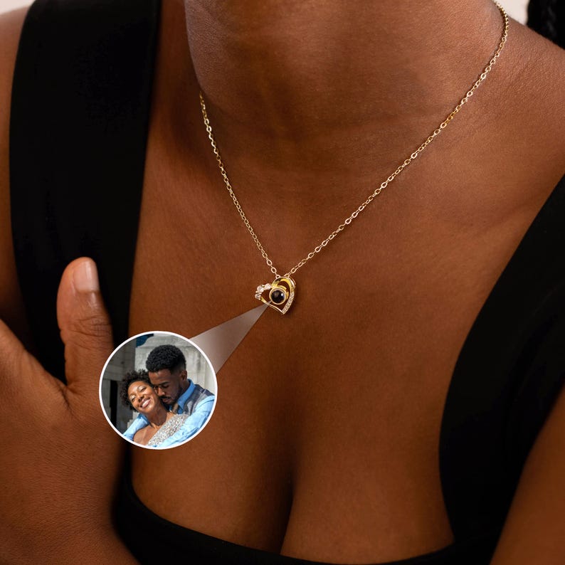 Peut inclure: Un collier pendentif en forme de c&oelig;ur dor&eacute; avec une petite pierre centrale ronde et sombre. Le pendentif est entour&eacute; de petites pierres claires. Une petite image ronde d'un couple est projet&eacute;e.