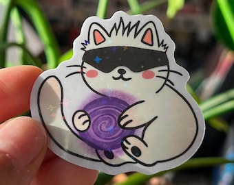 May include: A white cat sticker with a black mask and a purple orb. The cat has a cute design with pink cheeks and a star on the mask. The sticker is held in a hand in front of a background of green plants.