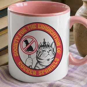 May include: A white ceramic mug with a pink interior and handle. The front features a graphic of a cat wearing a crown, holding a sign with a silhouette of a dog inside a red prohibition circle. The text reads Society for the Eradication of Cocker Spaniels.