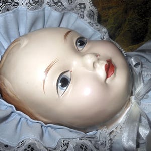 May include: A porcelain doll with painted features, wearing a blue and white lace bonnet and a blue and white dress.
