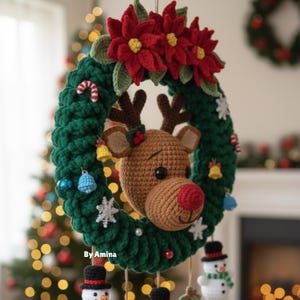 Op de afbeelding: Een handgemaakte kerstkrans met een gehaakte rendierkop, rode neus en geweien. De groene krans is versierd met rode poinsettia's, sneeuwvlokken en kleine ornamenten. De tekst "By Amina" is zichtbaar.