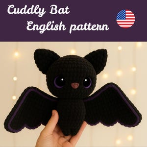 Op de afbeelding: Een zwarte gehaakte vleermuis met grote paars omrande ogen, een roze neus en paarse vleugelaccenten. De tekst "Cuddly Bat English pattern" staat erboven, met een Amerikaanse vlag. De vleermuis wordt in een hand gehouden.