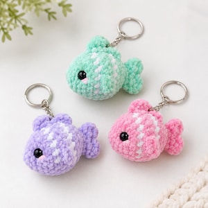 May include: Three crocheted fish keychains in mint green, lavender, and pink. Each plush fish features a textured yarn body, a white vertical stripe, black plastic eyes, and a silver-toned metal keyring attached to the top fin with a chain.