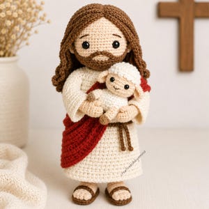 Puede incluir: Una muñeca de ganchillo que representa una figura con pelo largo castaño y barba, vestida con una túnica color crema con una faja roja y sandalias marrones. La figura sostiene un pequeño cordero de ganchillo blanco. El texto creativekidsbyamina es visible.
