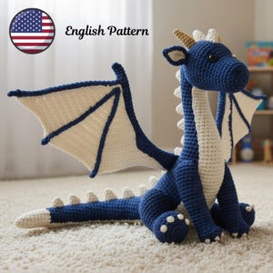 Puede incluir: Un dragón de ganchillo azul y crema hecho a mano con grandes alas, cuernos y una larga cola. El dragón está sentado sobre una alfombra color crema. El texto "English Pattern" es visible en la imagen.